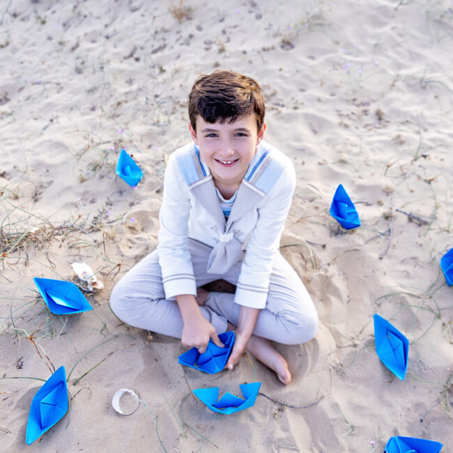 Niño de comunión jugando en la playa rodeado de barquitos de papel azul.