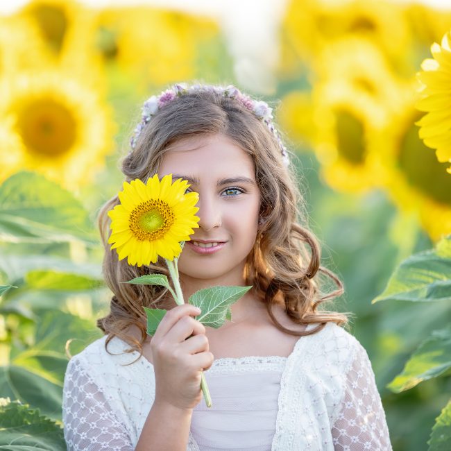 First Communion portrait of a girl holding a sunflower over one eye, surrounded by sunflowers