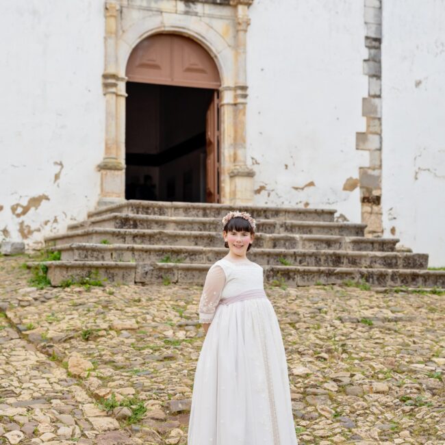 Isabel com vestido de primeira comunhão diante de um portal histórico em pedra no Algarve, Portugal